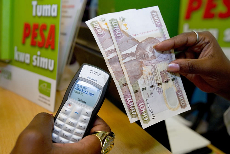 Person holding Kenyan shillings and a phone beside an M-Pesa sign